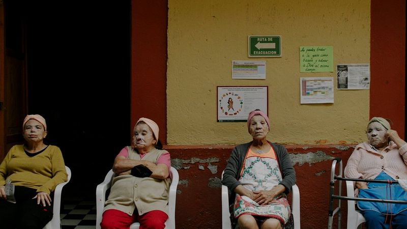 Four older women with beauty facial masks covering their faces sit in a line in front of a yellow and red wall, in this still from a documentary short by filmmaker Irene Baqué.