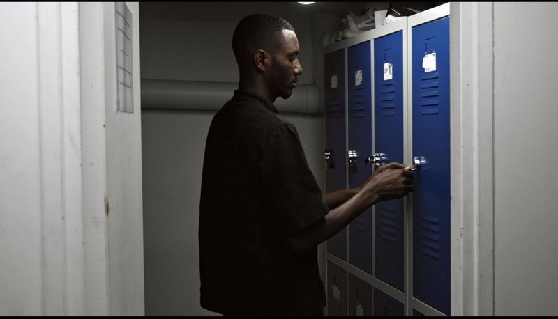 In a small room, a young man wearing a black shirt opens a blue locker, in this still from Camille Millerand's documentary short, The Invisibles, made with help from the Canon Video Grant. 