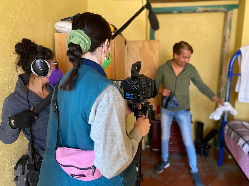 Two women, one holding a camera and one holding a mic, film a man in a small bedroom, placing clothes from a wardrobe onto a low bed.