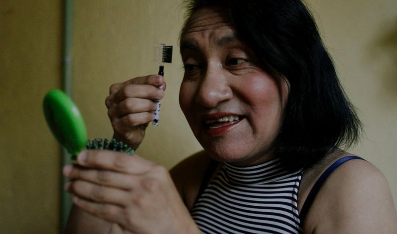 An older woman wearing a blue top and scarf holds up a piece of fabric to the side of the shot in this still from a documentary short by filmmaker Irene Baqué.