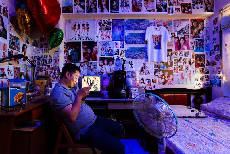 A man sits in his bedroom, looking at his phone and smiling. The room's walls are covered in images of female celebrities.
