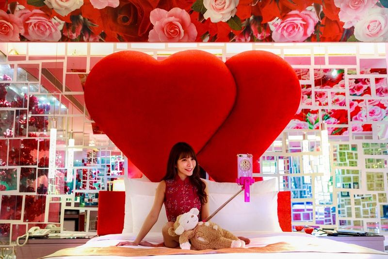 A young woman sits on a white bed, holding a long selfie stick with her phone on the end. There is a large display of two red hearts behind her.