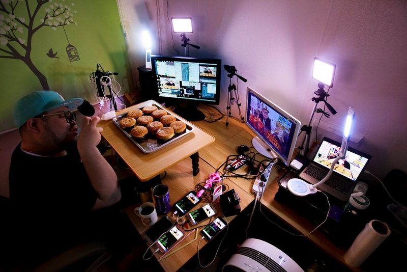 A man sits at a desk with a raised tray full of burgers in front of him. Also on the desk are two monitors, a laptop and several phones.