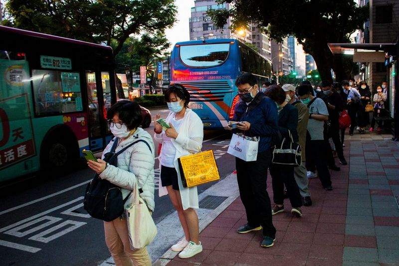 A queue of people stand at a bus stop, holding shopping. They are all wearing masks and looking at their phones.