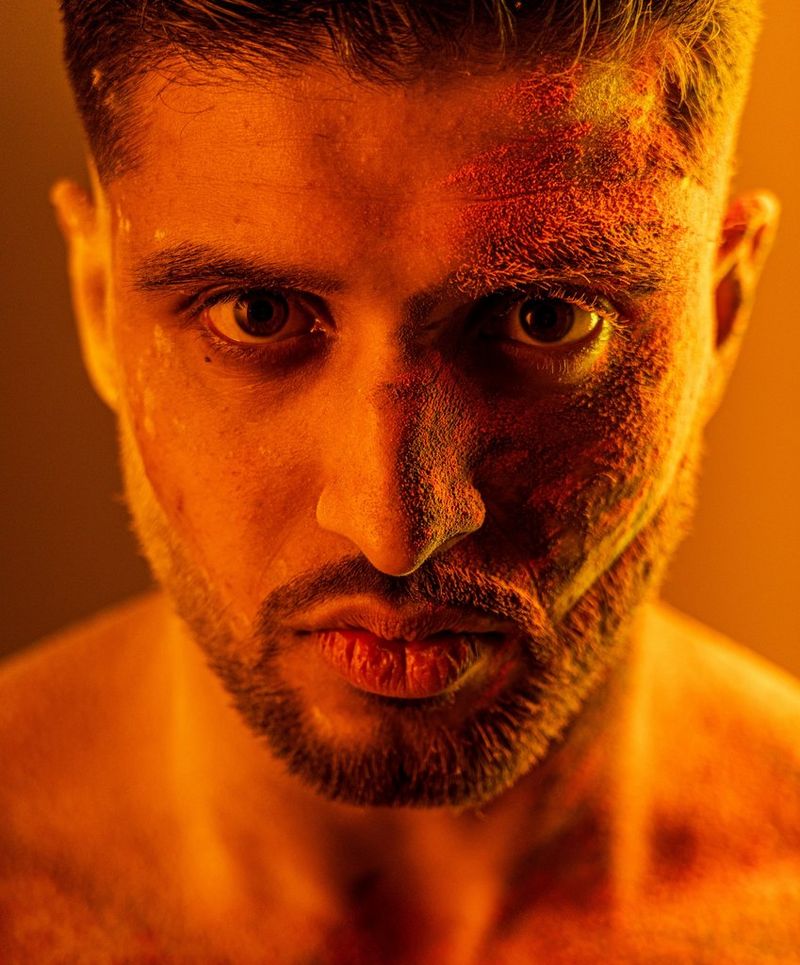 A close-up of a man's face, with an orange tint to the whole picture. He has a short, neat moustache and beard and coloured powder over one half of his face.