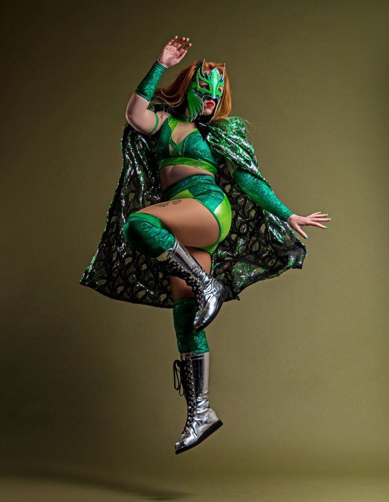 A female wrestler in a glittery green costume, cape and mask and sparkly silver boots poses as she jumps in the air.