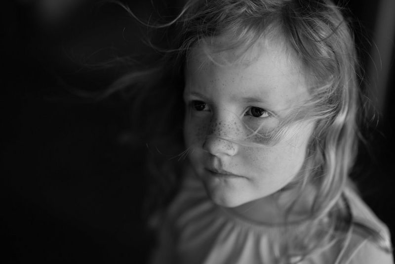 A black and white photograph of a young girl, facing to the side of the camera. A slight breeze is blowing strands of hair across her face and to the side.
