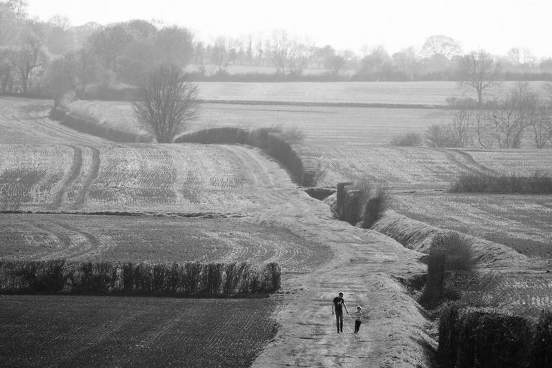 Shot from a distance, a black and white photograph of a man and a young boy holding hands as they walk along the edge of a field.
