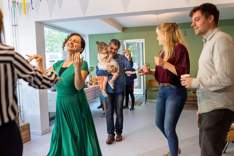Five adults in a living room dance and smile, one of them holding a child in a party dress. Partially obscured in the background are more children and a buffet table.