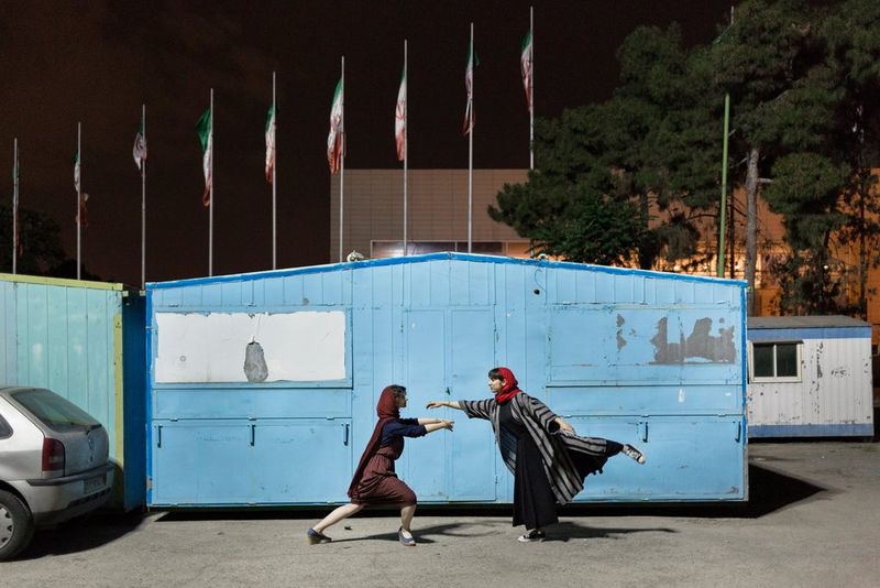 Twee danseressen in gewaden en een hoofddoek staan op het punt elkaar te omhelzen voor een blauwe hut, met een rij vlaggenmasten op de achtergrond.