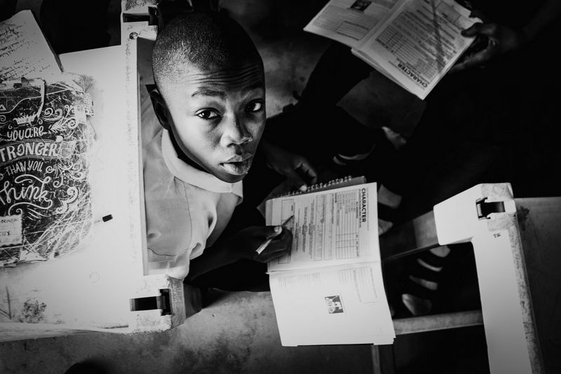 Een zwart-witfoto van een jongen die op de vloer van een klaslokaal zit en een formulier op zijn schoot invult. Hij kijkt omhoog naar de camera.