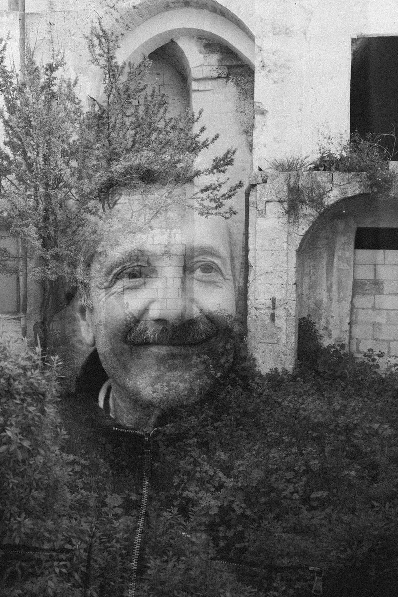 A black and white double exposure of a man with a moustache and a stone archway covered in wild plants.