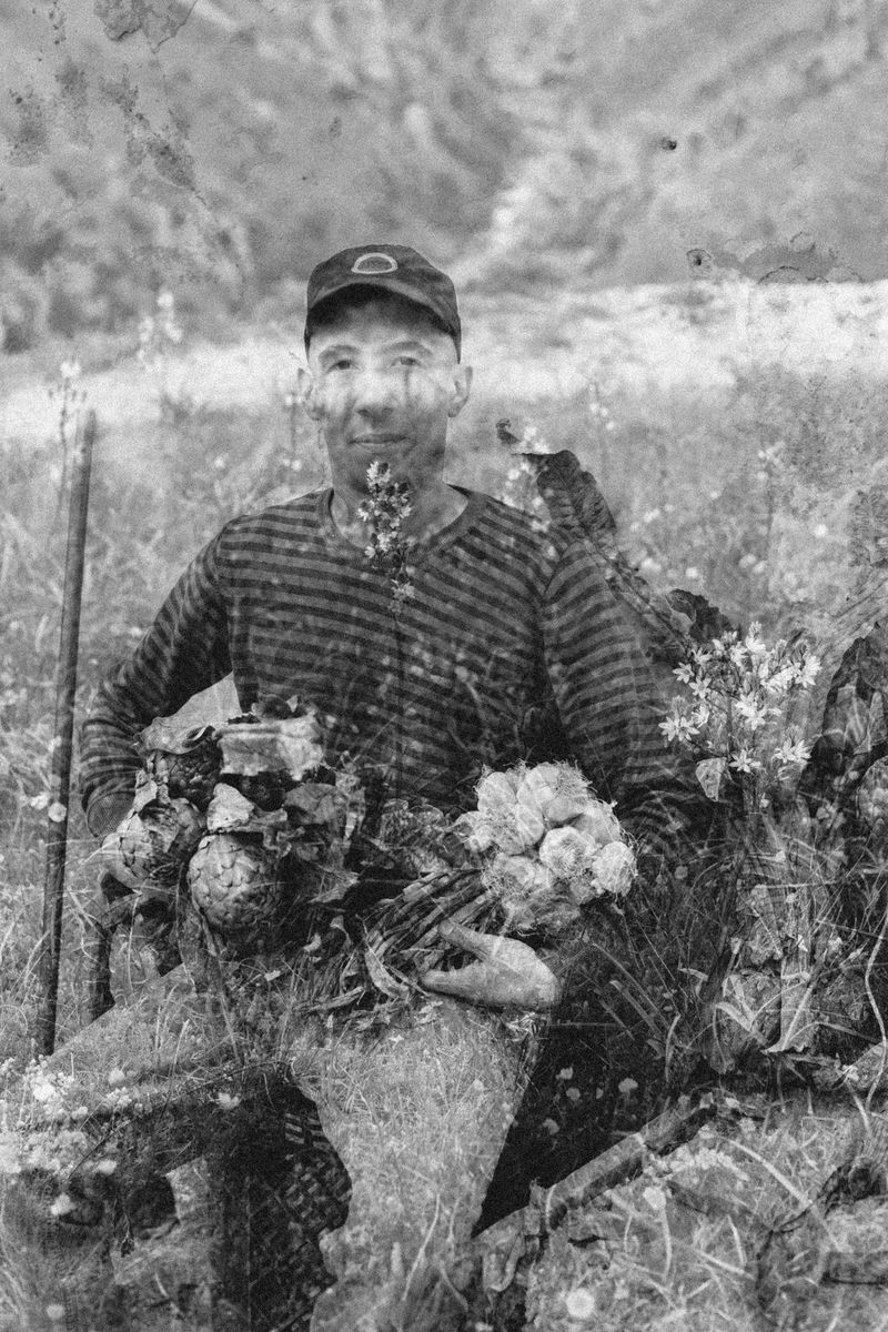 A black and white double exposure of a seated man holding bunches of flowers and a field of wildflowers.