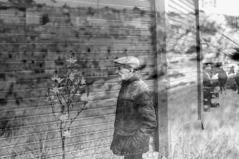 A black and white double exposure of a man in a flat cap walking beside a wall and a close-up of a small tree in bloom.