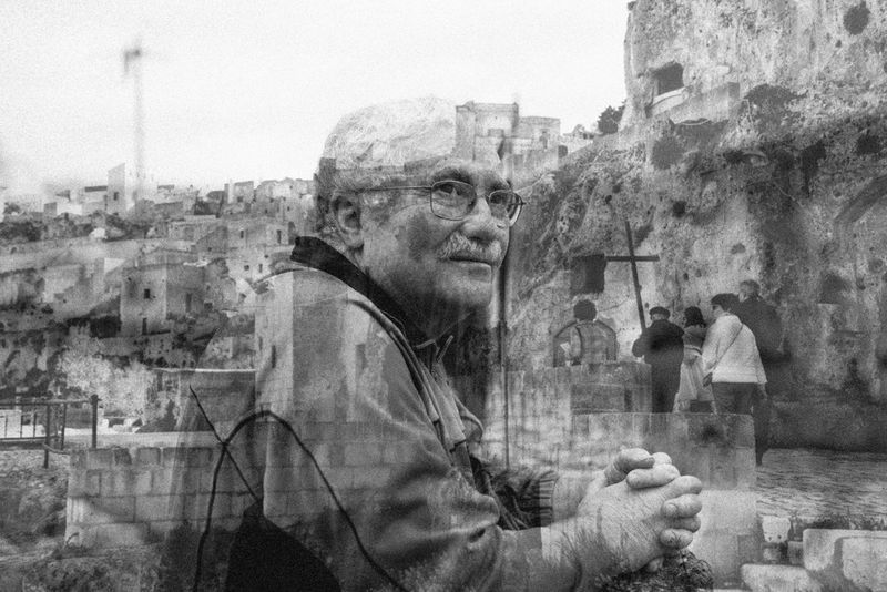 A black and white double exposure of a man with glasses leaning against a wall and a religious procession through stone streets.