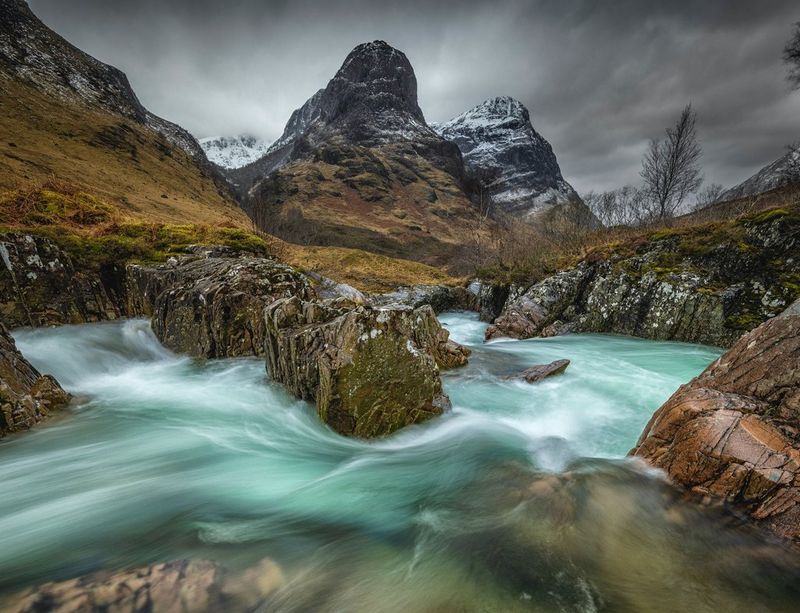 A one-second exposure of a fast flowing river surrounded by mountains. The long exposure has blurred the movement of the water.
