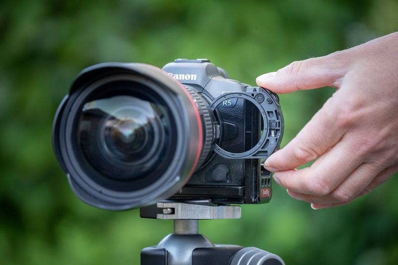 A camera with large lens sits on a tripod. A hand reaches in to place a filter in the mount adapter at the back of the lens.