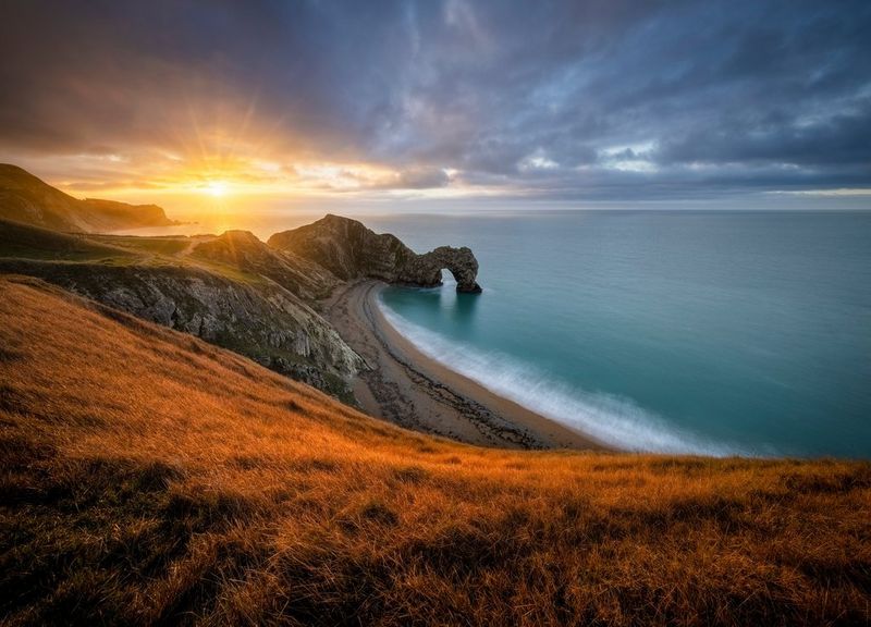 The sun rises from the sea, lighting up a dramatic rocky coastline and grassy landscape.