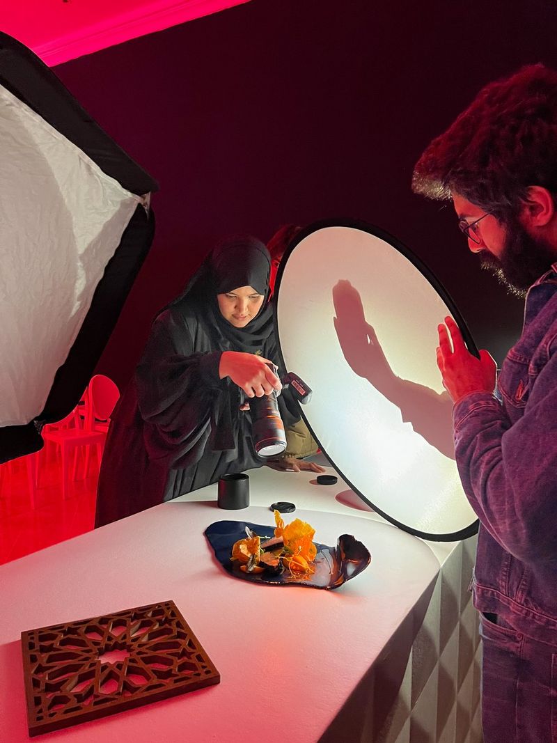 A man using his hand behind a light reflector to cast a shadow in the frame as food photographer Yasmin Albatoul captures a food composition in the studio with a Canon camera. Shot on Canon.