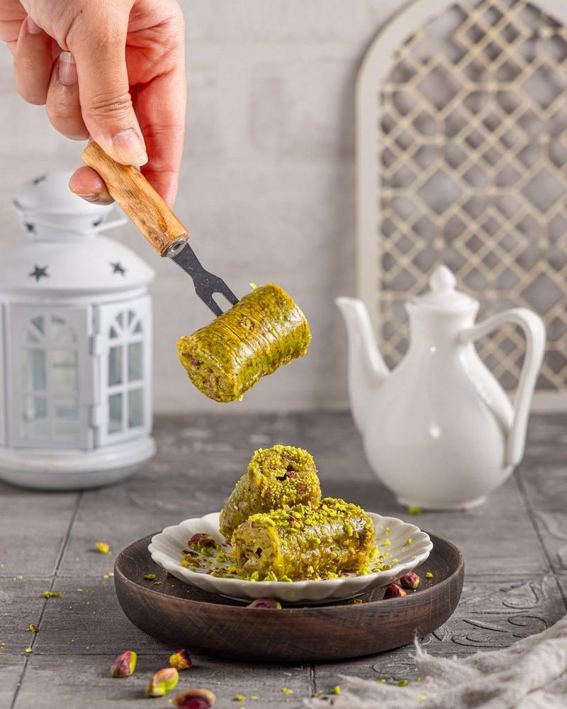 A hand holding a skewer pierced with a piece of pistachio baklava over a plate while syrup drips down from above. Shot on Canon.