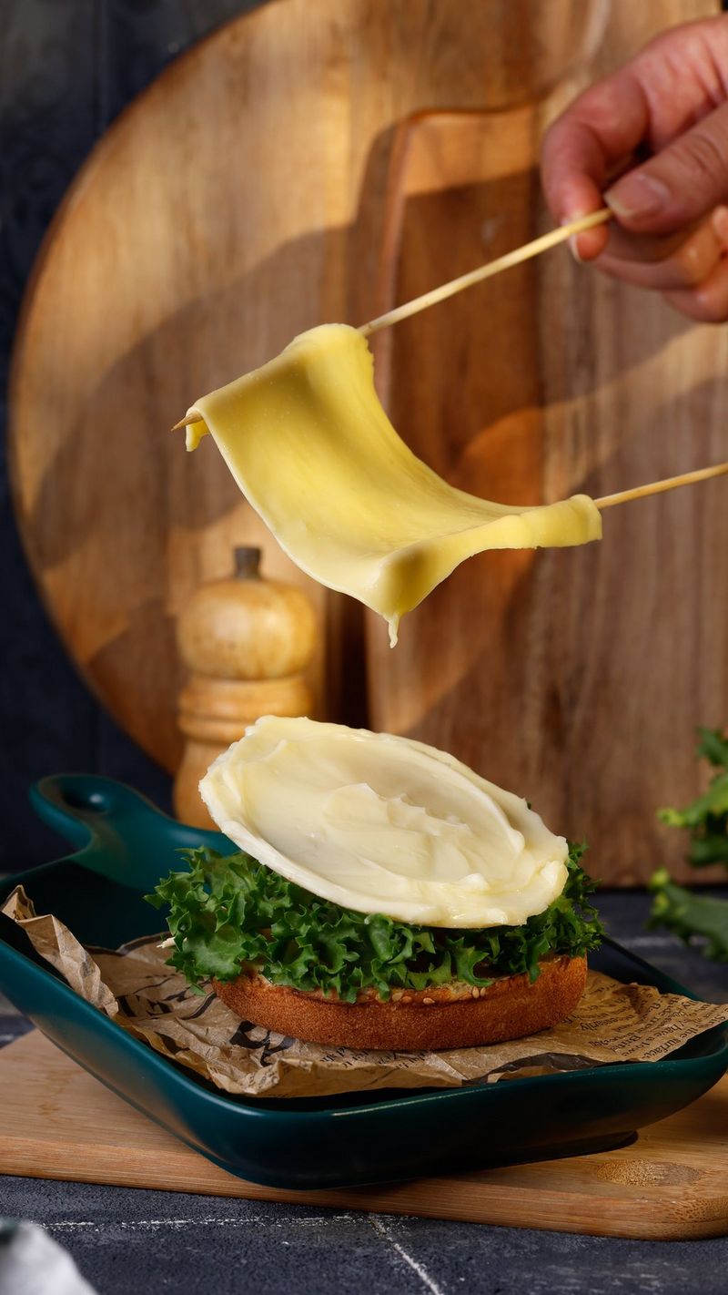 A hand suspending a piece of melted cheese with two wooden sticks above a bun topped with green leaves and fake white sauce. Shot on Canon.