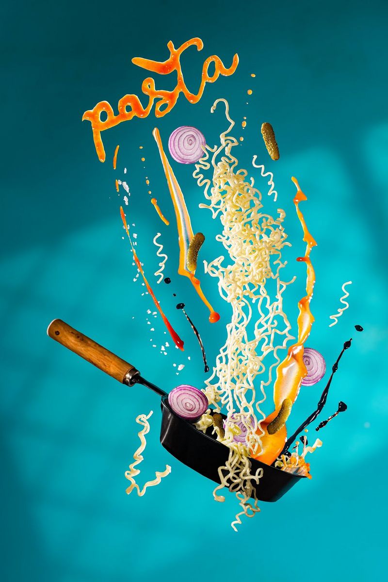 Yellow curly noodles, sauce and sliced onions falling into a pan suspended in the air in front of a teal background with the word pasta framing the top of the food.