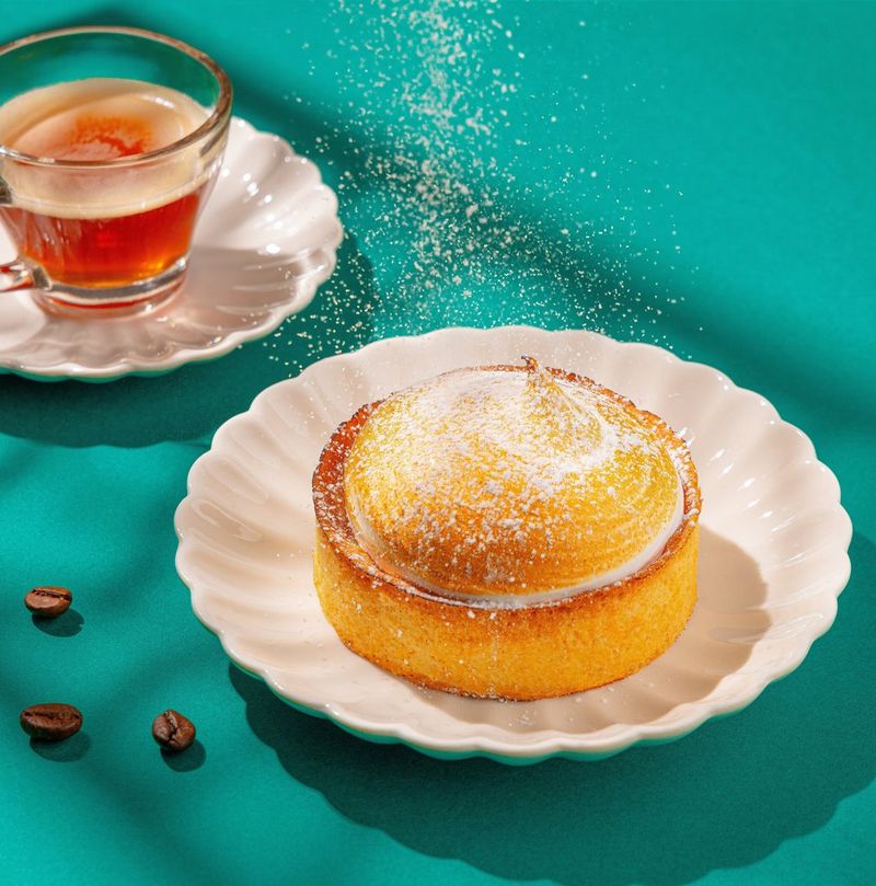 A white scalloped plate holding a small lemon meringue pie next to a glass of cream tea laid on a green background scattered with coffee beans. Shot on Canon.