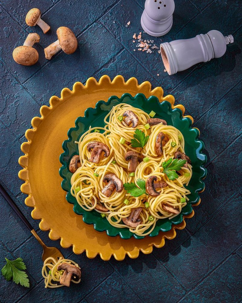 Overhead view of a plate of small nests of spaghetti topped with slices of mushrooms and green leaves on a dark green plate with a colour chart sitting in the bottom right corner. Shot on Canon.