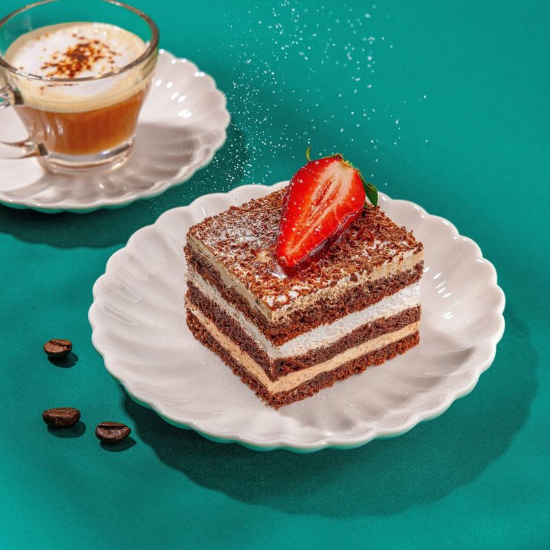 A light dusting of icing sugar falls onto a square of coffee cake topped with a slice of strawberry on a white scalloped plate, with a glass cup of cream tea on the side, laid on a green background with scattered coffee beans. Shot on Canon.