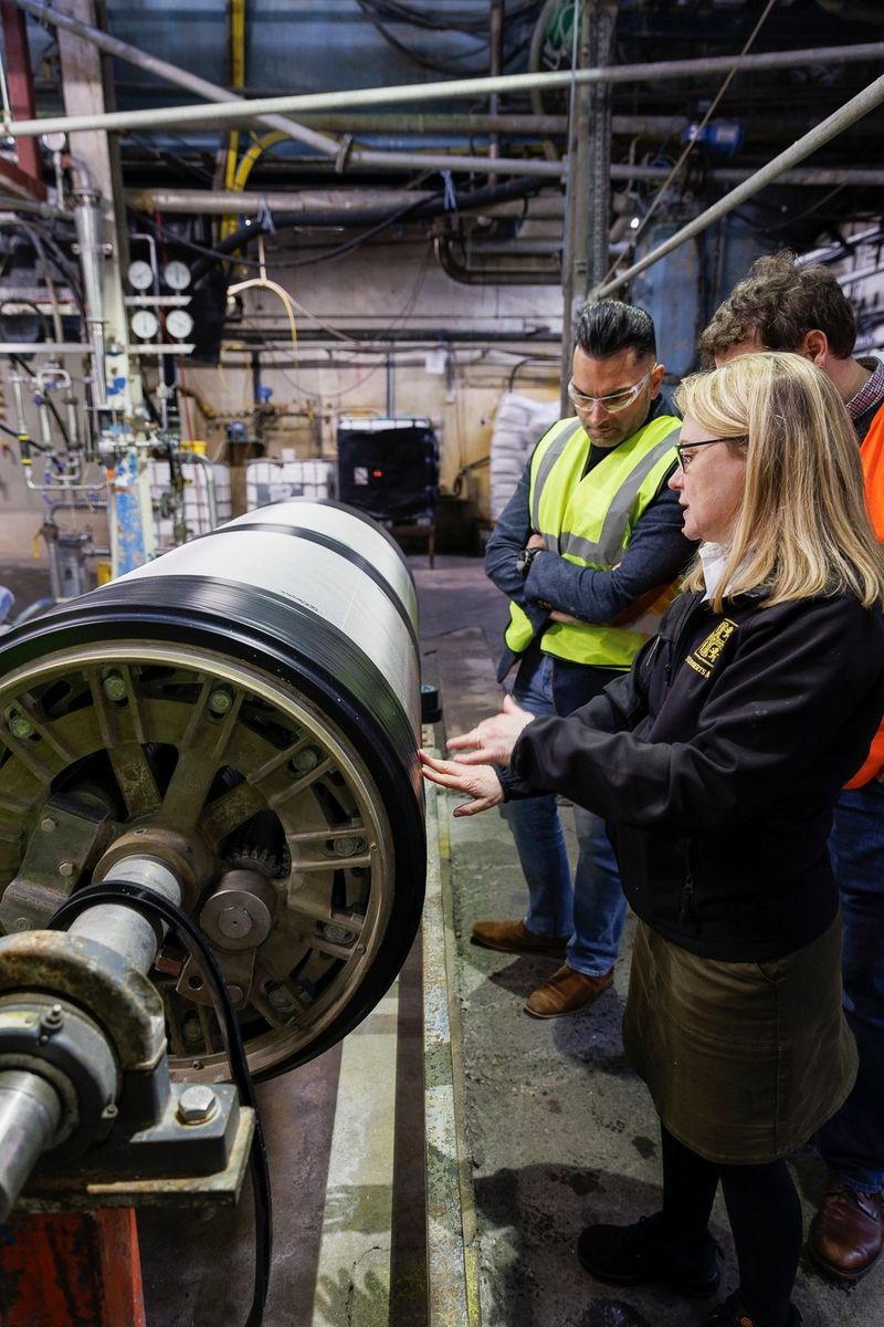 Catherine Frood, employée de St Cuthberts Mill et notre guide pour la journée, présente la machine Cylinder Mould utilisée dans le processus de fabrication du papier, à côté de deux hommes portant un gilet réfléchissant et des lunettes de sécurité. Photo prise avec un appareil Canon.