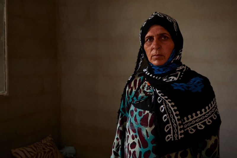 Женщина-беженка из Ирака в платье с орнаментом и платке на голове стоит перед темным фоном.