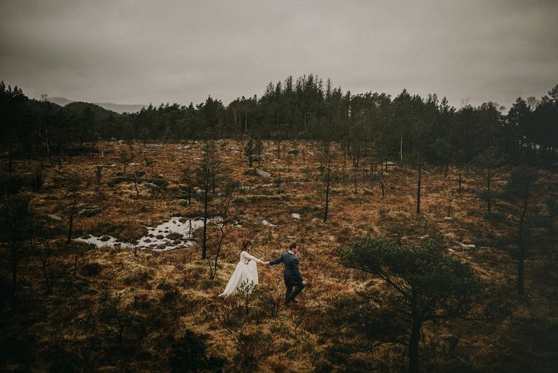 Una coppia di sposi cammina attraverso un campo punteggiato di alberi ai margini di una foresta.