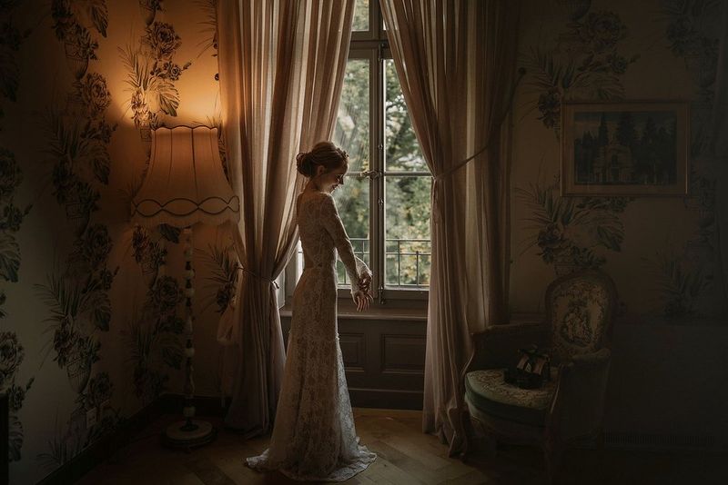 Una sposa in piedi vicino ad una finestra in una lussuosa camera d'hotel. Le sue scarpe sono poggiate sulla sedia al lato.