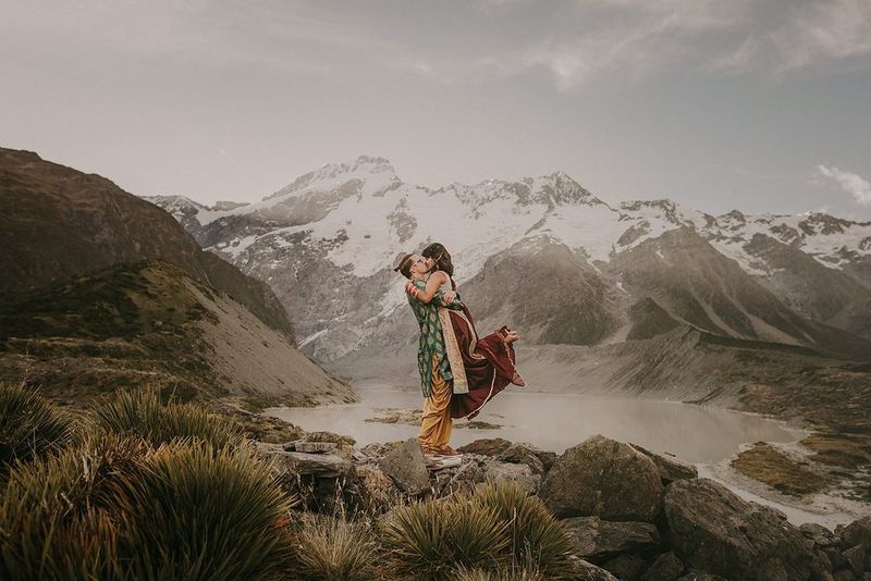 Una coppia di sposi si bacia su una collina con una serie di montagne innevate sullo sfondo.