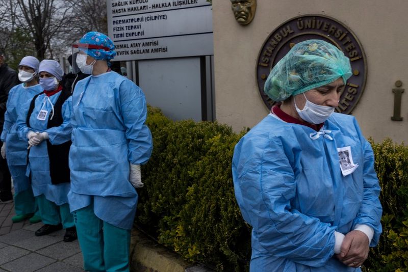 Four medical professionals wearing masks, plastic hats and blue plastic overalls stand outside a hospital. 