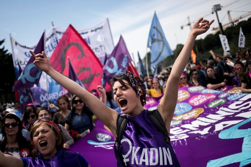 Amid a large, flag-waving crowd, a woman wearing a headscarf and a bright purple top shouts and holds her arms aloft. 