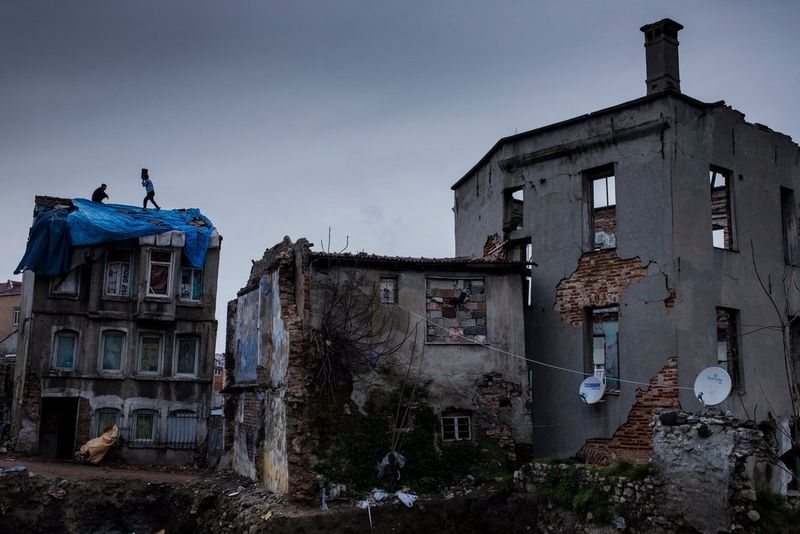 Two young men can be seen in the distance repairing the roof of a dilapidated building. In the foreground is another equally broken-down building.