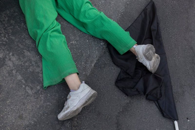 The legs of a young woman lying on concrete. She is wearing green trousers and white trainers and her right foot is resting on a black flag.