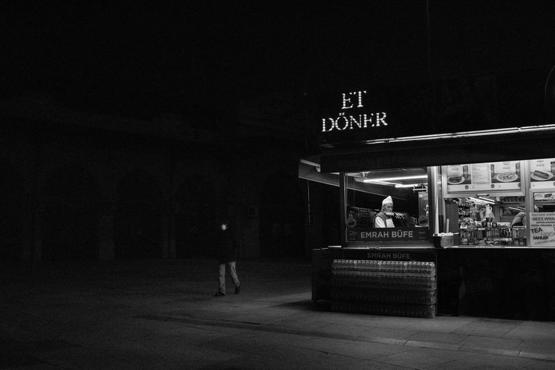 A black-and-white image of a man wearing a mask walking past a brightly lit but empty takeaway restaurant.