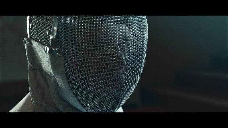 Un primo piano di uno schermidore che indossa una maschera da scherma con un intenso sguardo fisso, ripreso in Canon Log 3. L'immagine è stata sottoposta a colour grading in post-produzione.
