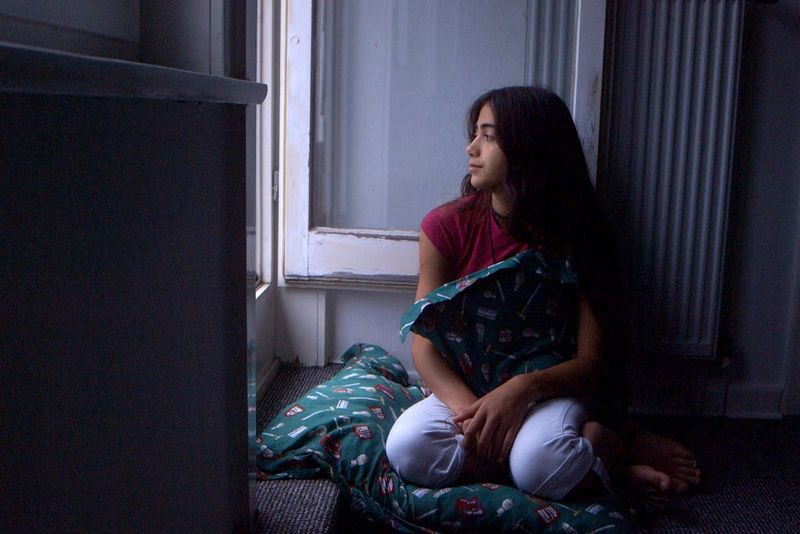 Une fille est agenouillée près d'une fenêtre ouverte et regarde dehors, tenant un oreiller à motifs entre ses mains.