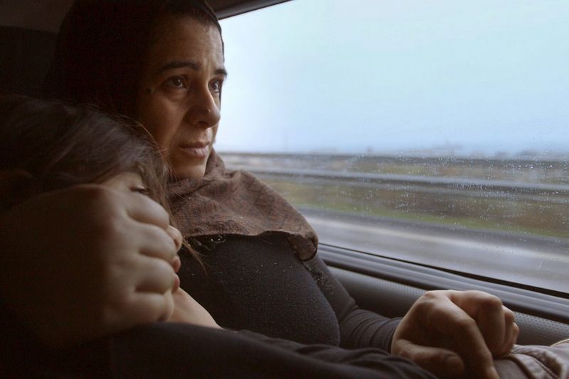 Assise sur le siège de la banquette arrière d'une voiture, une femme tient son jeune enfant dans ses bras. Par la fenêtre, on peut distinguer une autoroute pluvieuse.