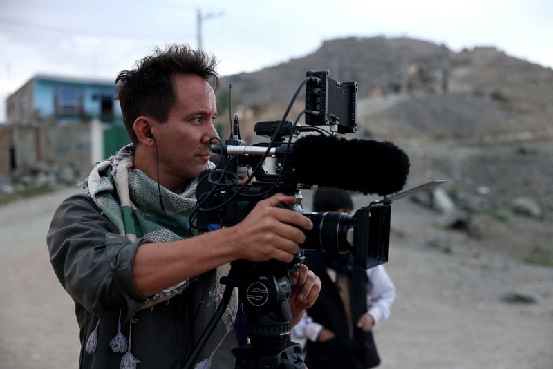 Un homme vêtu d'une veste kaki et d'une écharpe se tient derrière une caméra de cinéma Canon sur un trépied.