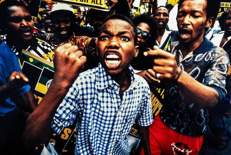 In een historische foto die Brent Stirton maakte met een Canon EOS-1 in Zuid-Afrika in 1994, steekt een boze jonge man in een geruit shirt zijn vuist omhoog, omgeven door een vurige menigte tijdens een protest.