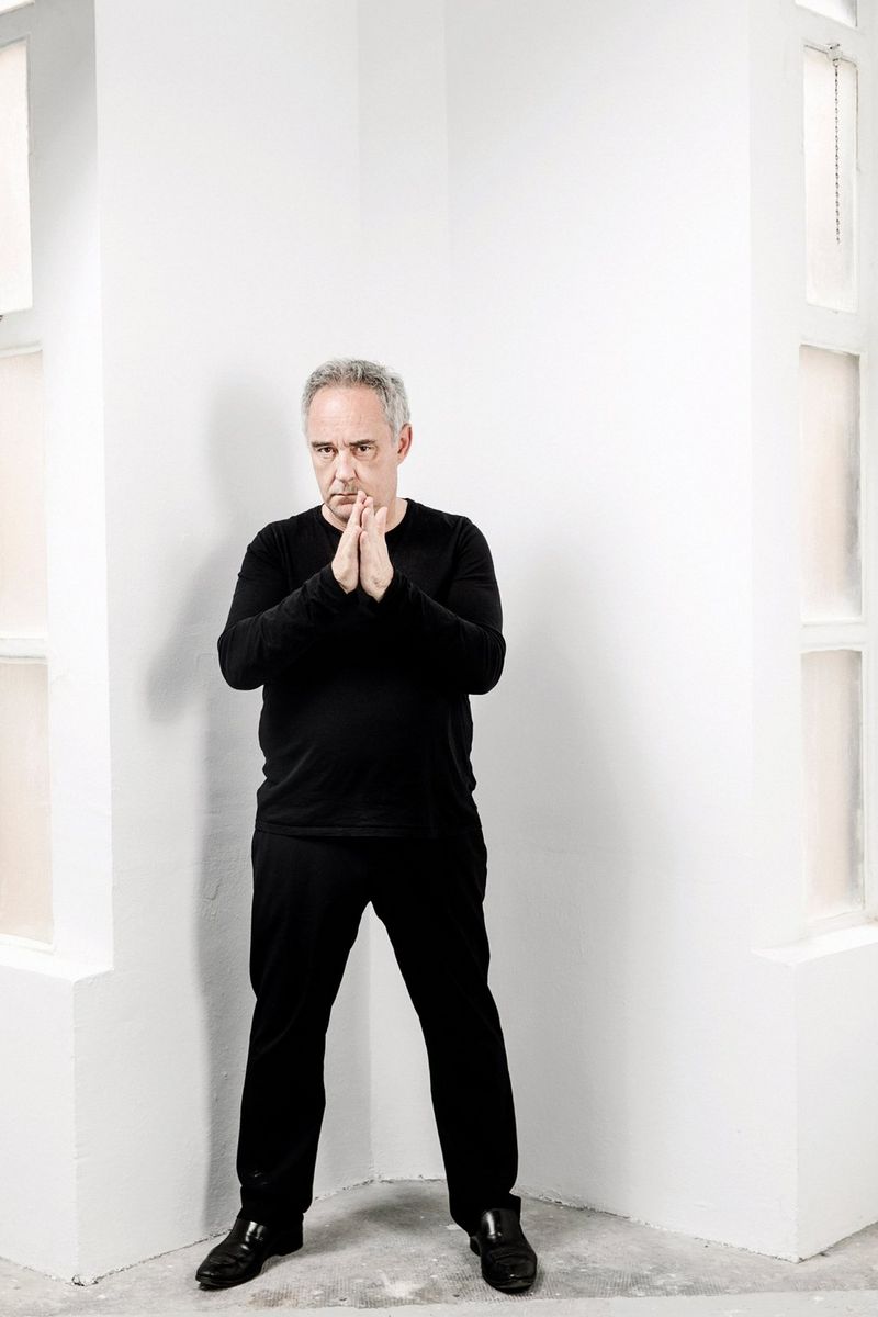 A portrait of Spanish Chef Ferran Adrià dressed in black and standing against a white wall.