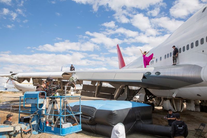 A cinematographer on a forklift films a dancer on the wing of a 747 in the desert.