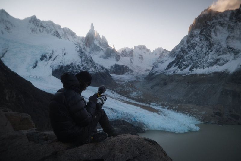 Filmmaker Daniel Ernst sits on a rock in front of a mountainous landscape, filming with the Canon EOS C50 that is fitted with 70-200mm lens and microphone.