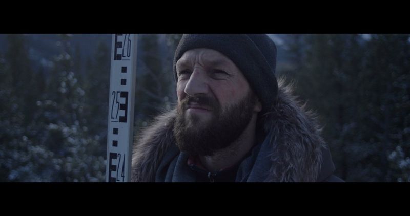 A still from a music video showing a bearded person, in a warm hat and coat, standing in a snowy setting next to a numbered stick.