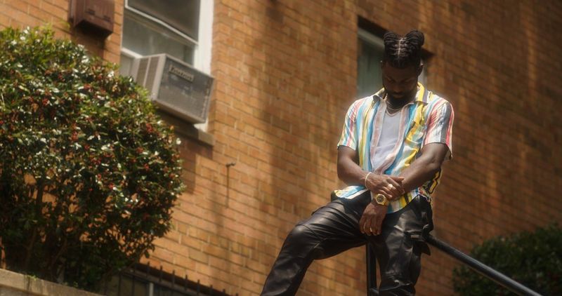 A man stands in front of a brick building, looking down and holding one wrist with the other hand.
