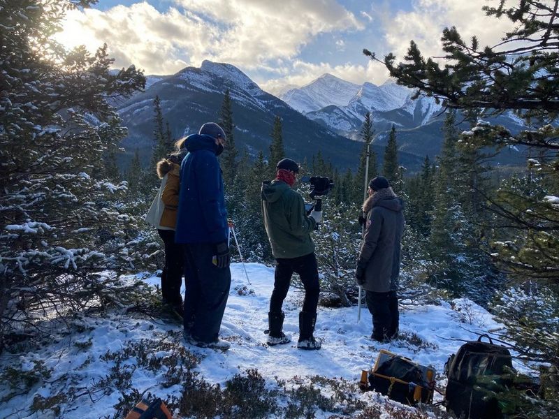 Four people, all dressed in warm winter clothing, filming in the snow. Behind them is a dramatic mountain range. 
